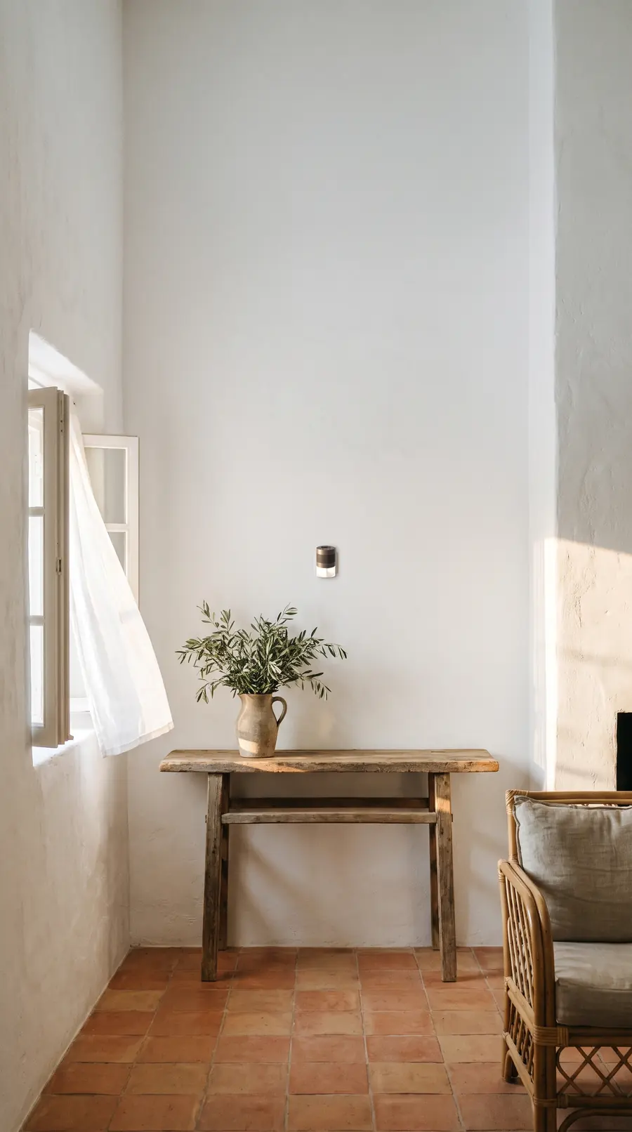 Pièce de maison de campagne : murs blancs chaulés, sol en tomettes, console rustique avec un pichet de branches d'olivier, fenêtre ouverte sur un voilage. Un diffuseur MOSK anthracite est fixé au mur au-dessus de la console.