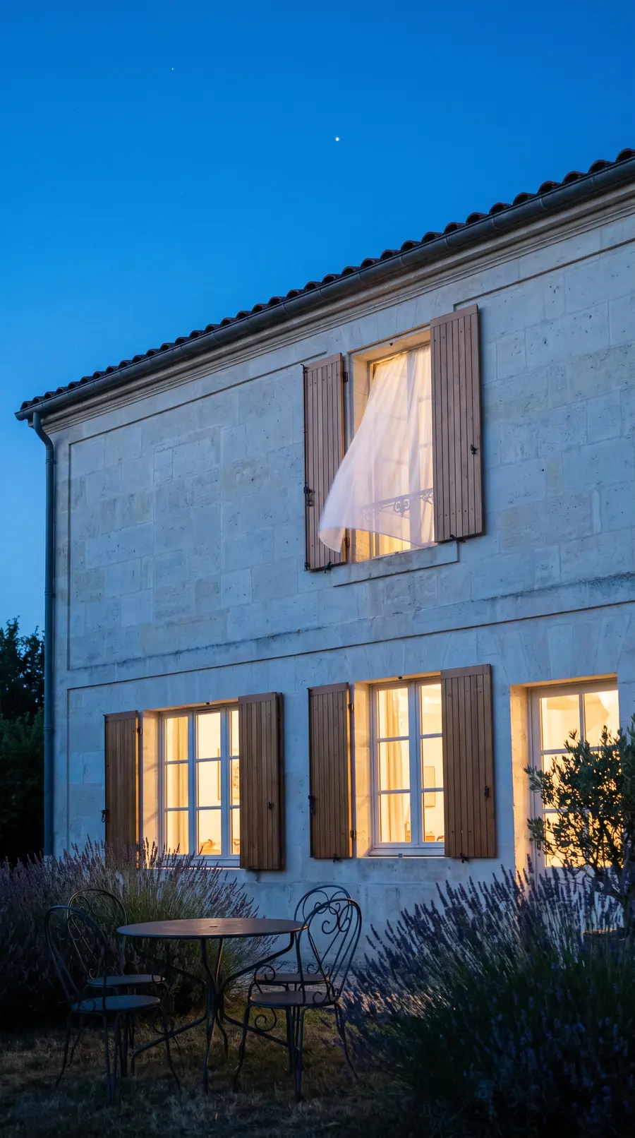 Une maison de famille en pierre à l'heure bleue, fenêtres éclairées, un voilage s'échappe d'une fenêtre ouverte à l'étage, lavande au premier plan.